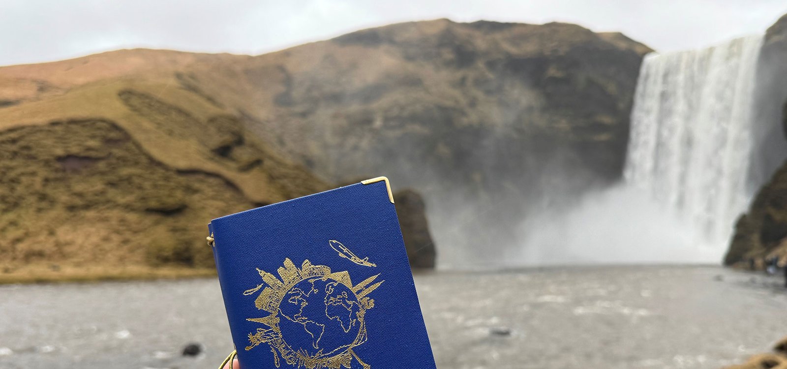 Travelfolio mit goldener Weltkartenverzierung wird vor einem Wasserfall in einer natürlichen Landschaft gehalten